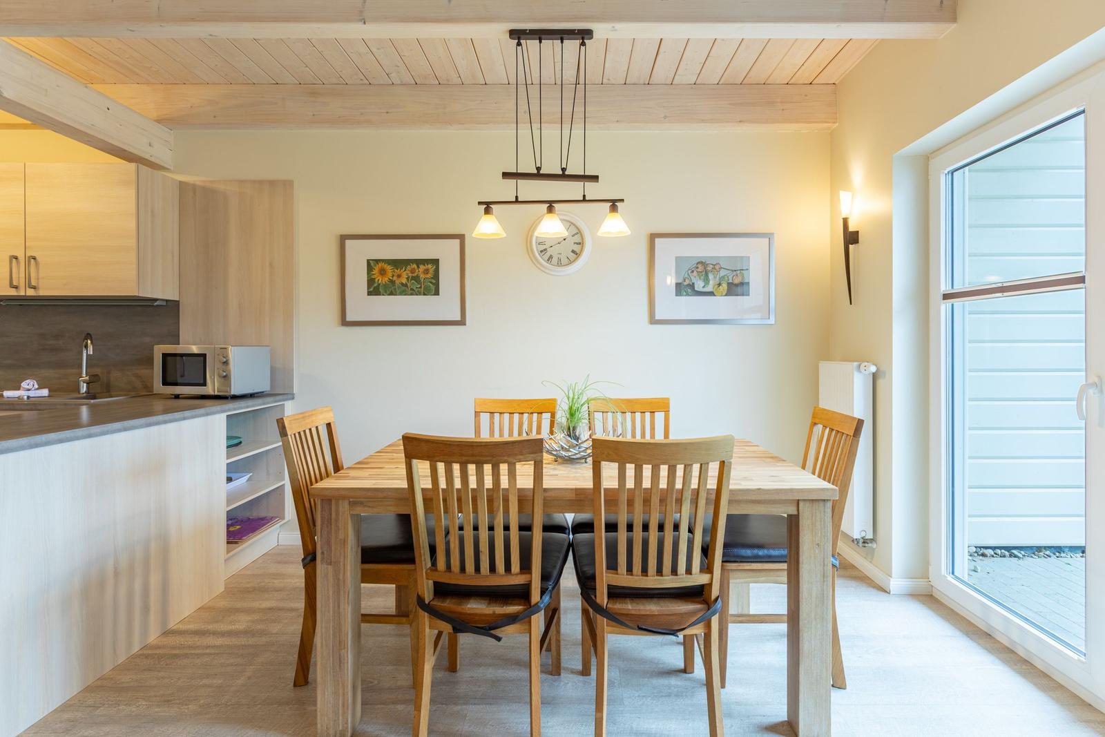 Esszimmer mit Holztisch, Stühlen und Küchenbereich. An der Wand hängen zwei Bilder und eine Uhr.