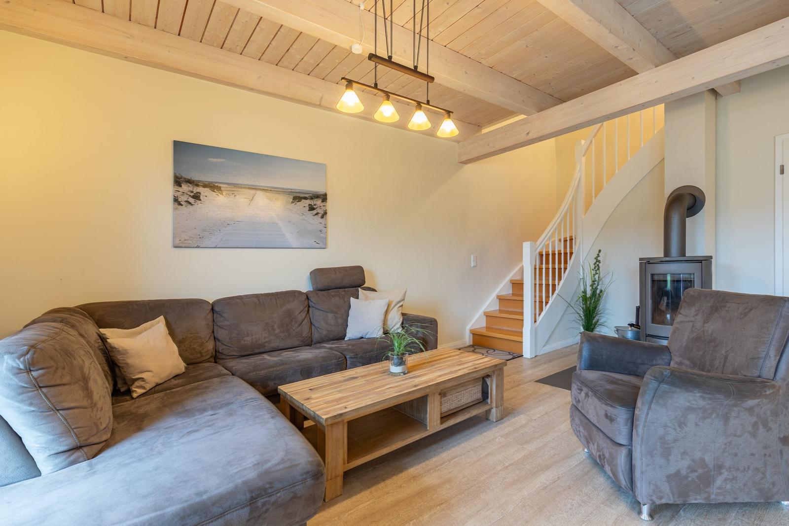 Wohnzimmer mit Sofa, Sessel, Couchtisch und Kaminofen. Holzdecke und Treppe sichtbar.