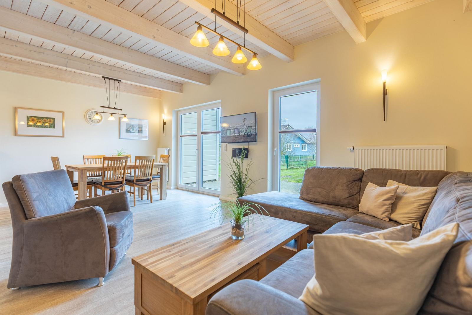 Wohn- und Essbereich mit Sofa, Sessel und Holztisch. Holzdecke und große Fenster.
