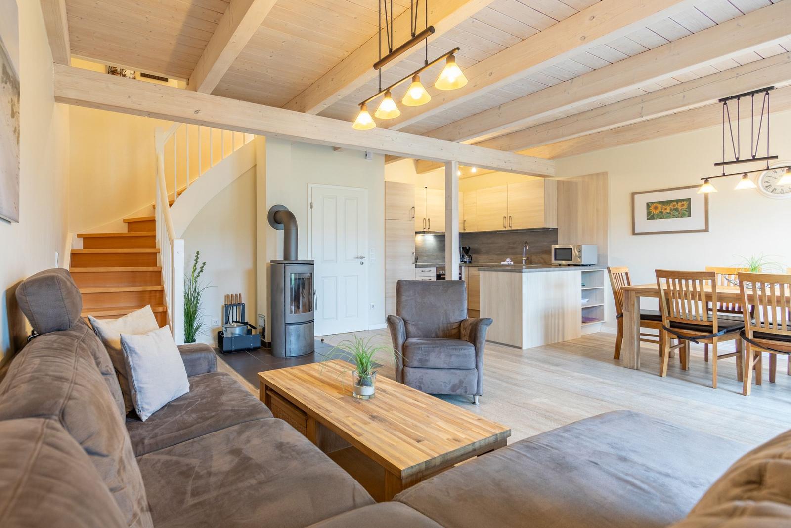 Wohnbereich mit Sofa, Sessel und Esstisch. Holzdecke und Kaminofen sichtbar.