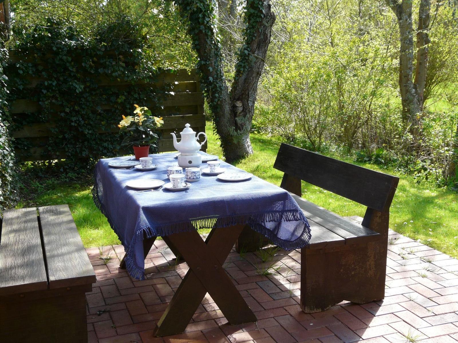 Holztisch mit Tischdecke und Teeservice auf Steinplatten im Garten.