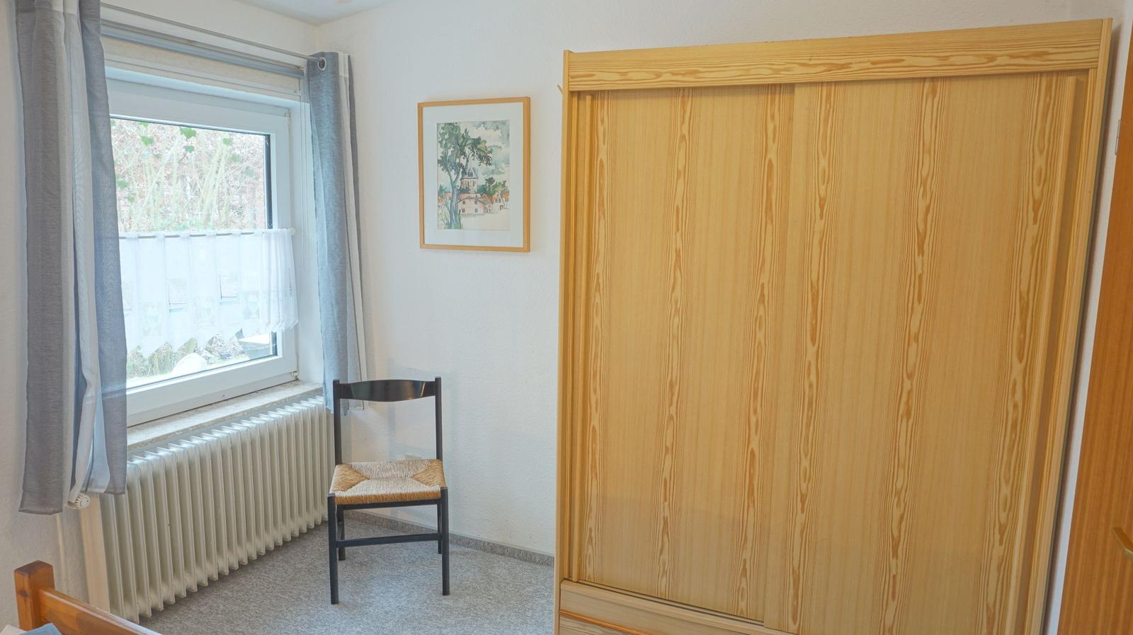 Ein Zimmer mit Fenster, Stuhl und Kleiderschrank.