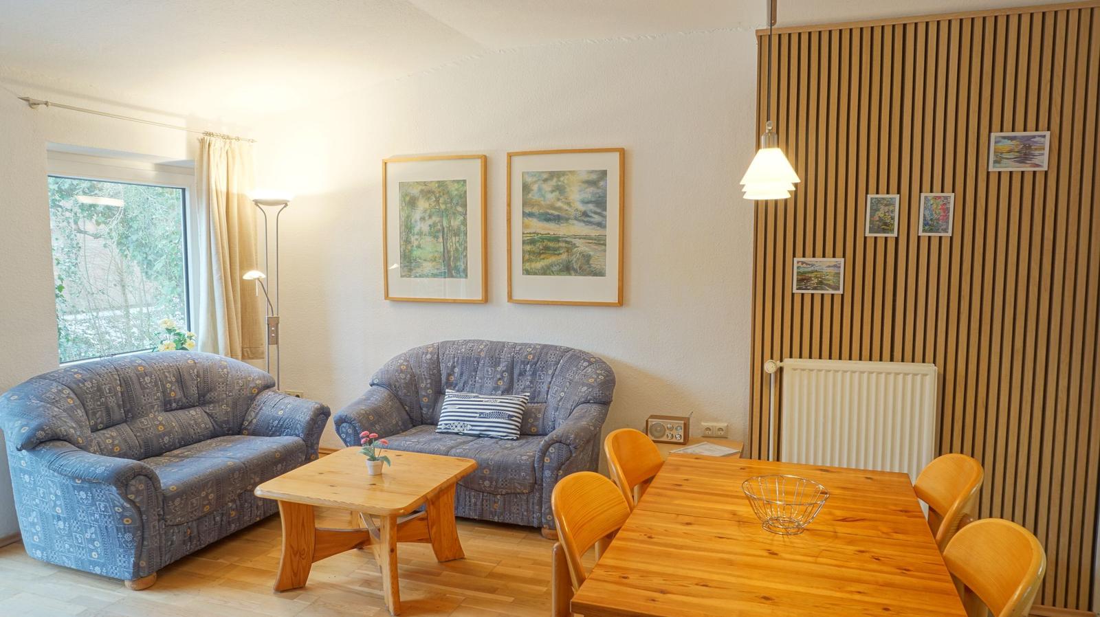 Wohn- und Essbereich mit Sofa, Tisch und Stühlen. Holzwand und Fenster sichtbar.