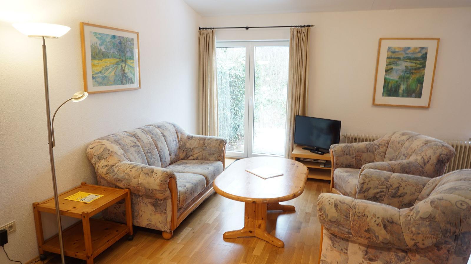 Wohnzimmer mit Sofa, Sesseln, Esstisch und TV. Fenster mit Vorhängen.