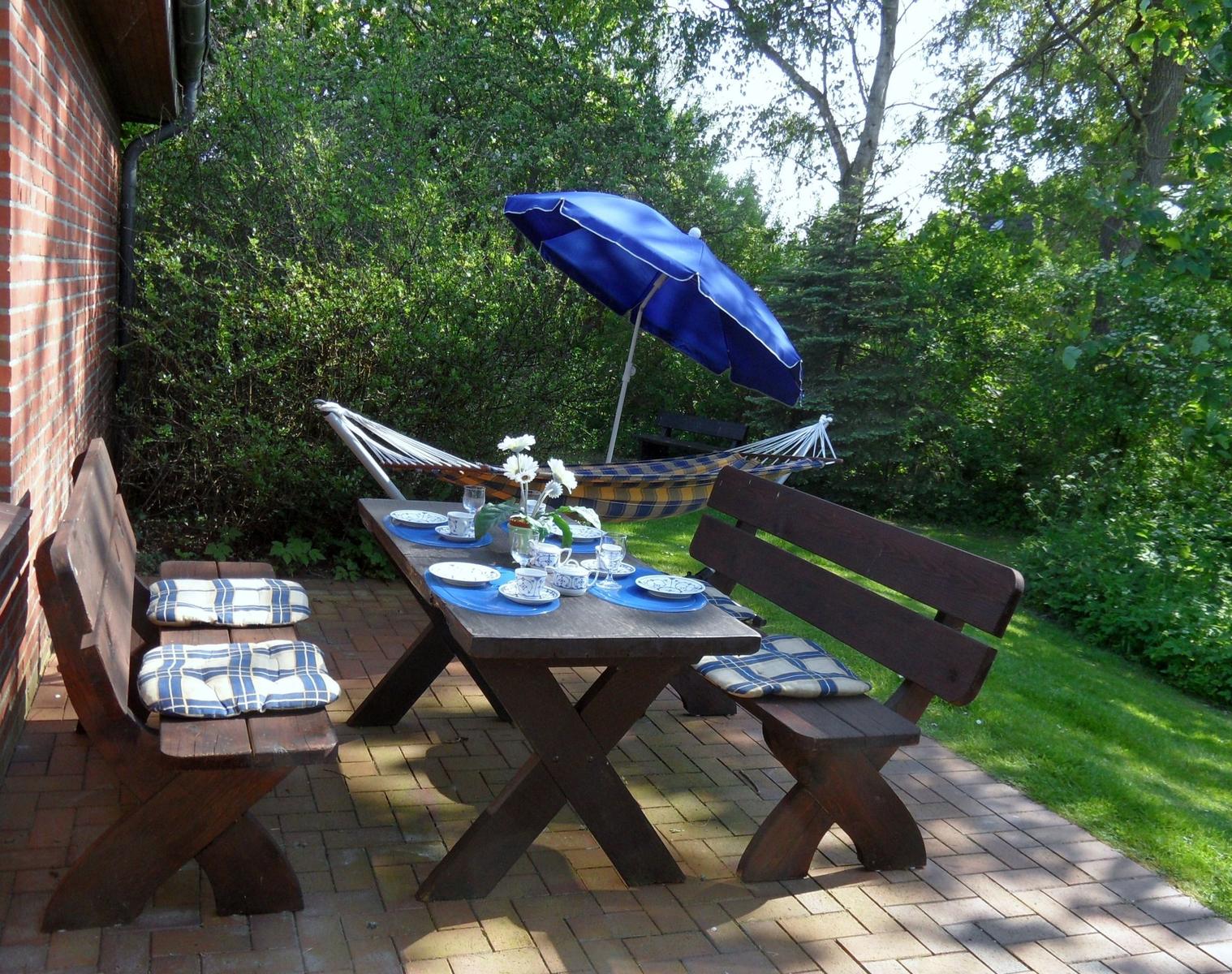 Holztisch mit Bänken, blauer Sonnenschirm und Hängemöbel im Garten.