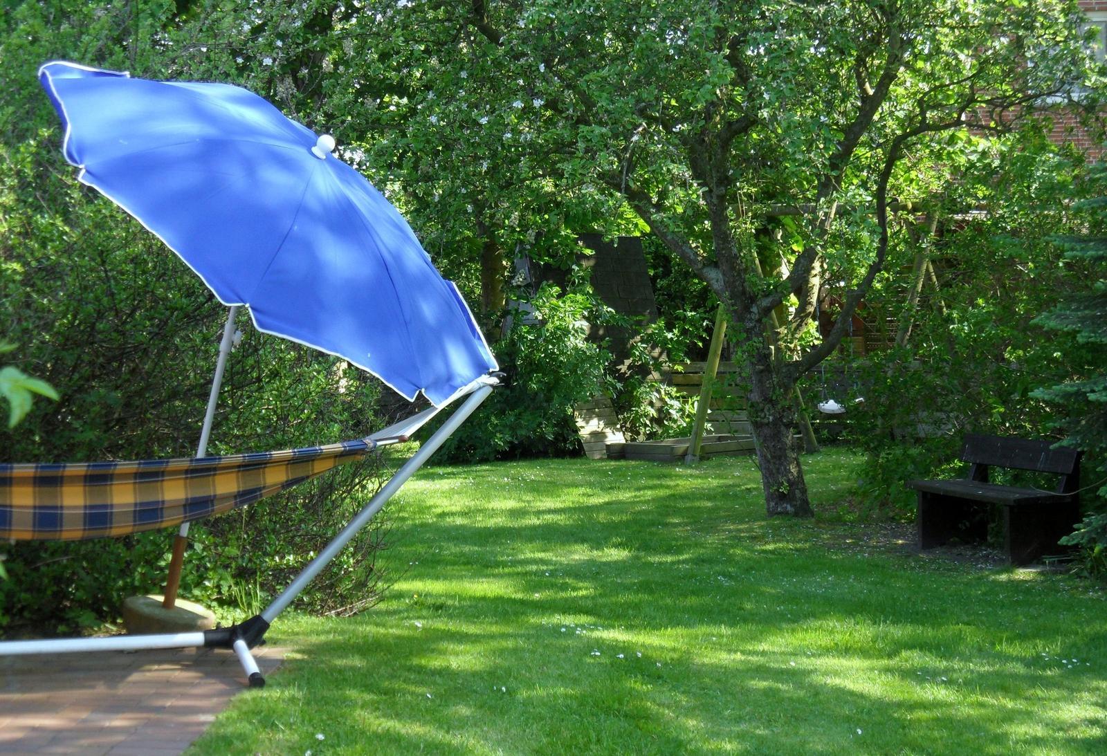 Garten mit Hängemöbel unter blauem Sonnenschirm, Holzbank und Bäumen.