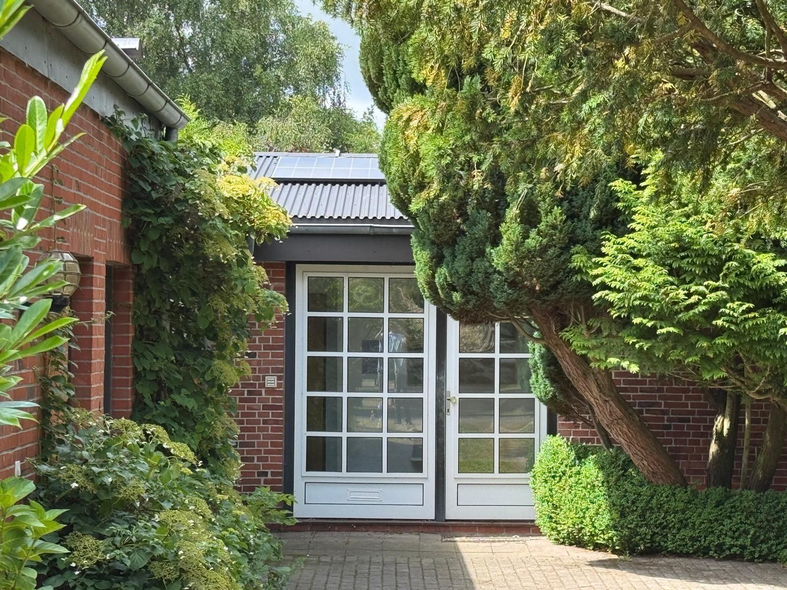 Eingangstür mit Glasfenstern, umgeben von grüner Vegetation und einem roten Ziegelbau.