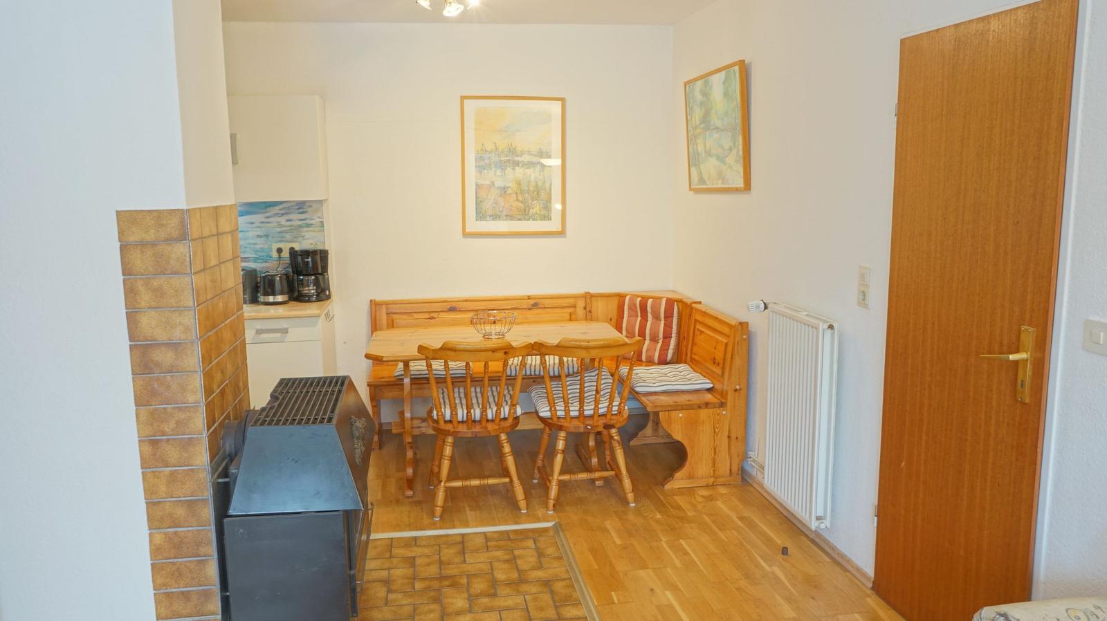 Essbereich mit Holztisch, Bänken und Stühlen. Küche und Kachelofen sichtbar.