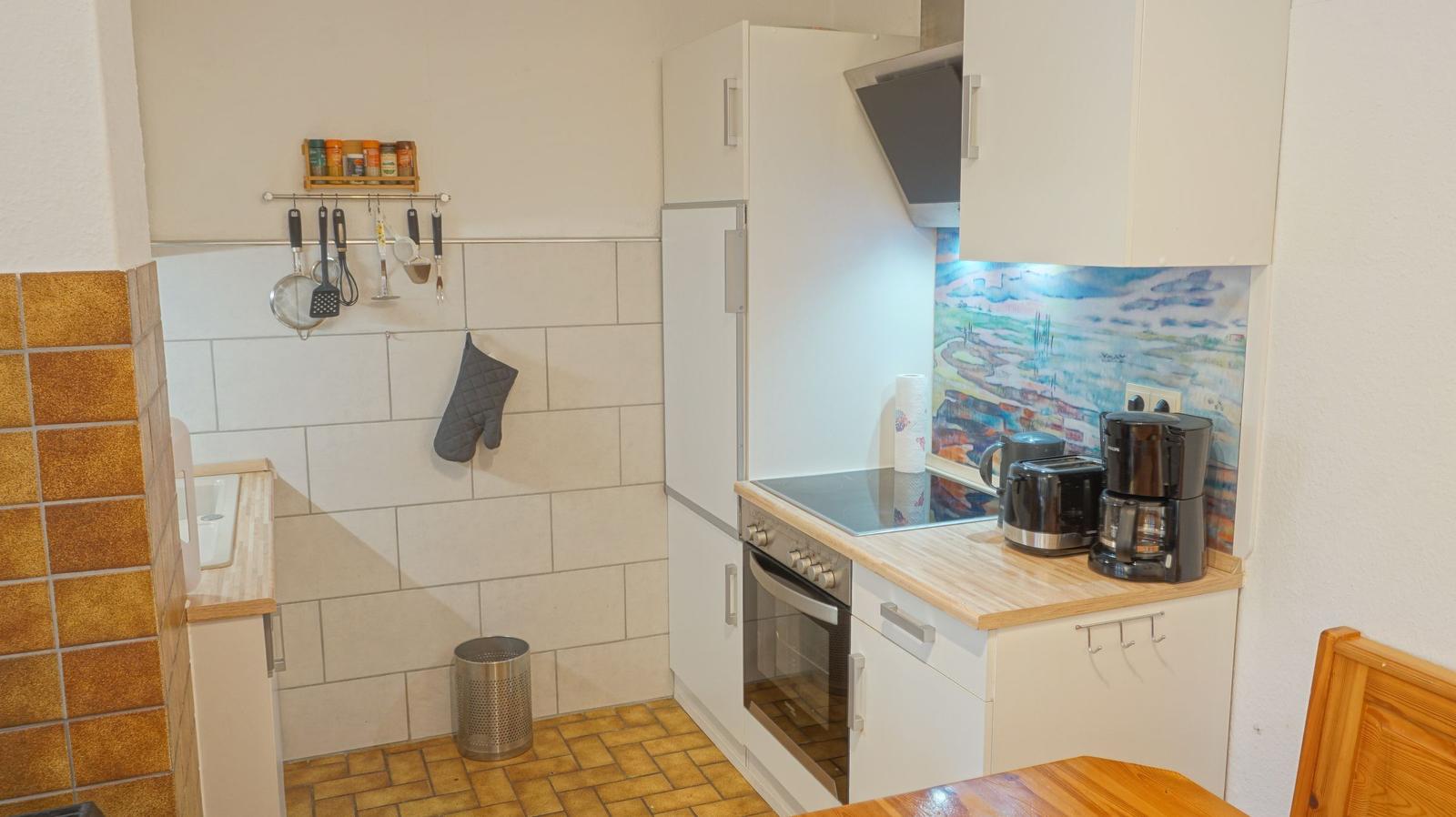 Küche mit weißen Schränken, Herd und Kühlschrank. Arbeitsfläche aus Holz. Wand mit Bild und Küchenutensilien.