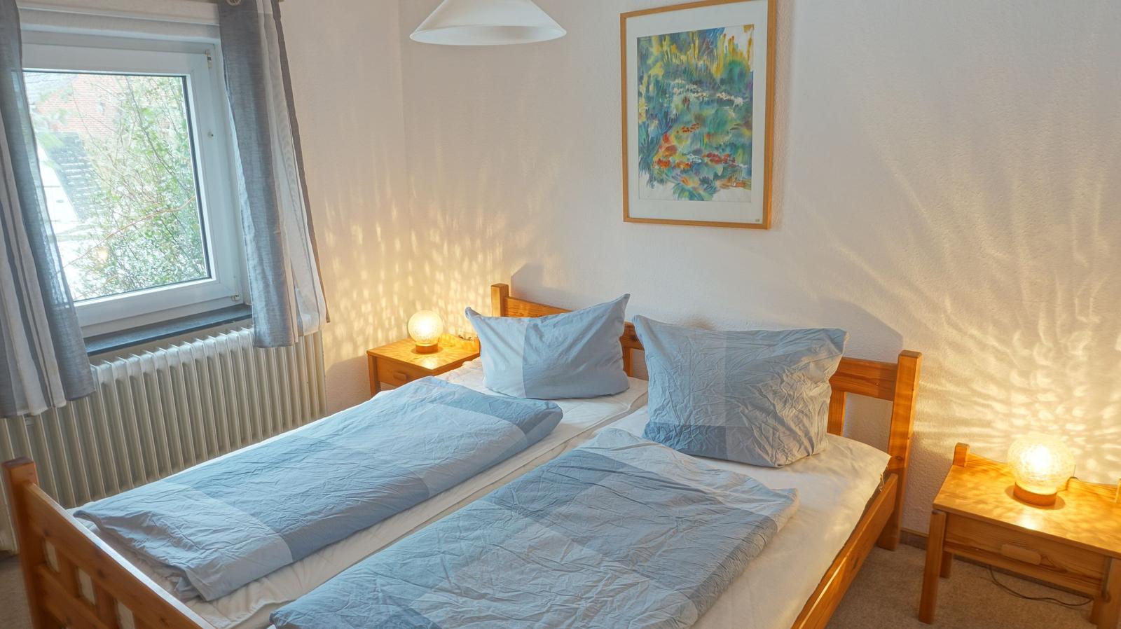 Zwei Einzelbetten mit blauen Bettwäschen und Holzrahmen. Fenster mit Vorhängen und Wandbild.