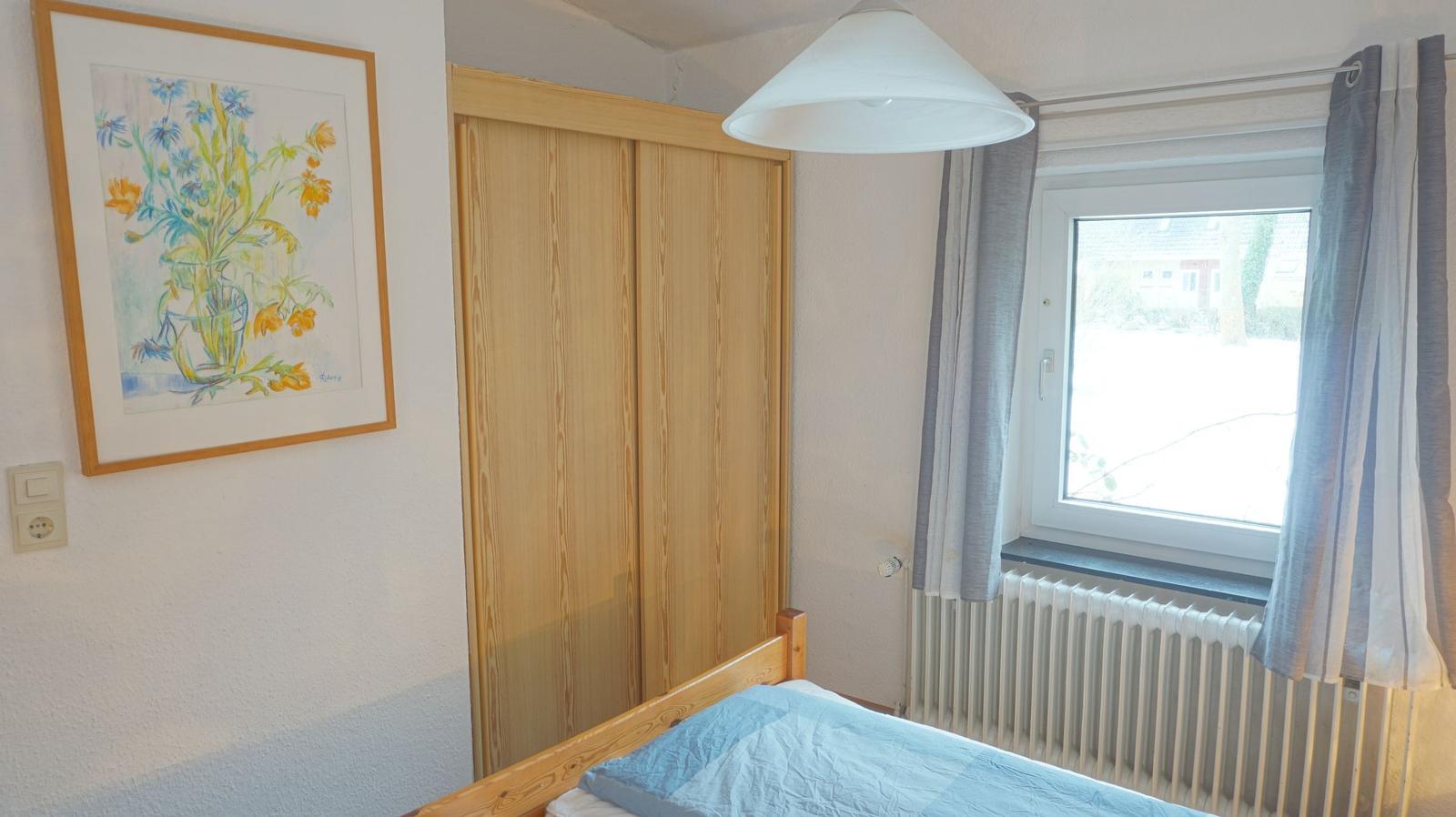 Schlafzimmer mit Bett, Schrank, Fenster und Wandbild.