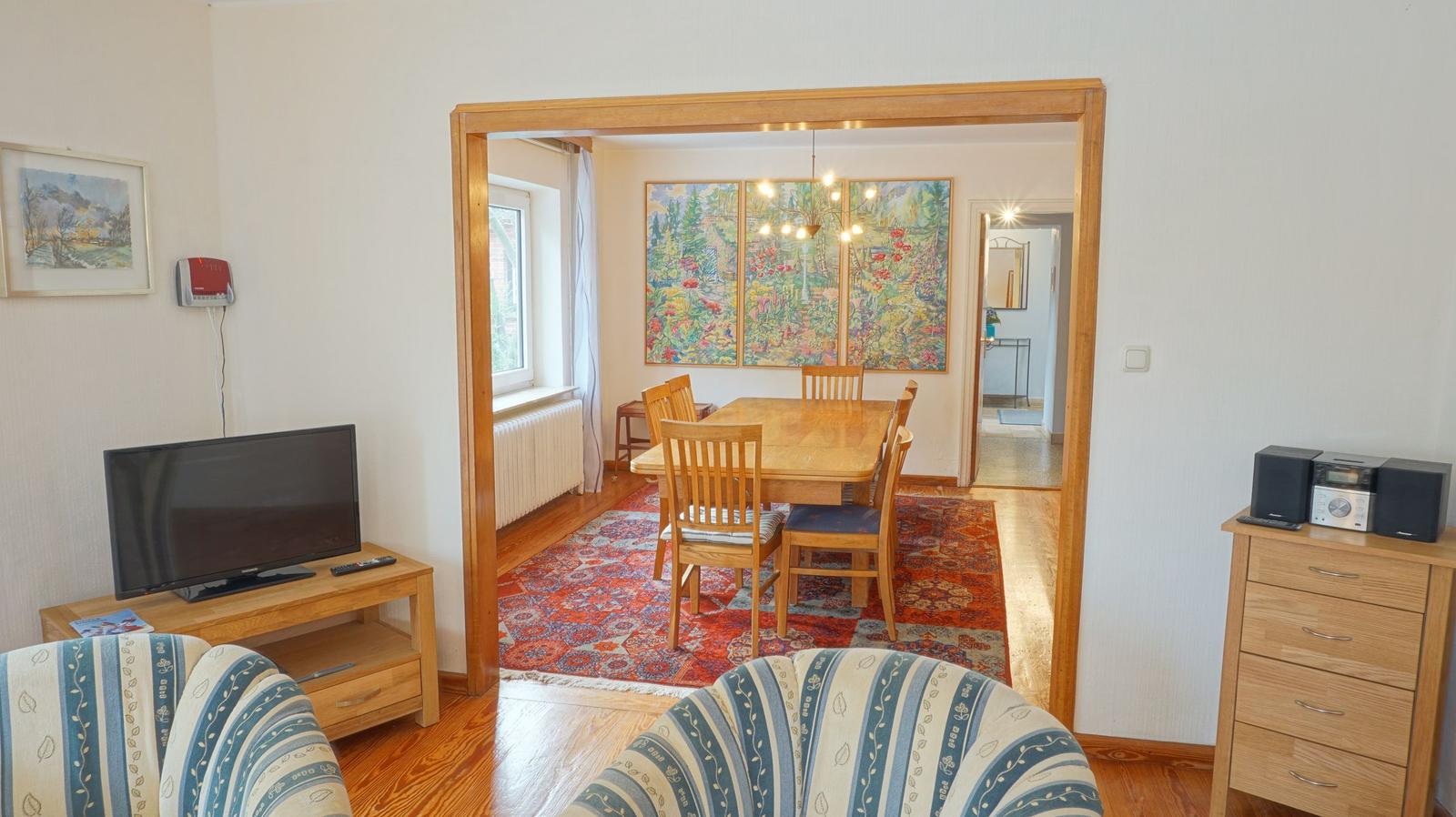 Wohnzimmer mit Fernseher und Sitzgelegenheiten. Durchgang zu Esszimmer mit Tisch, Stühlen und Wandbildern.