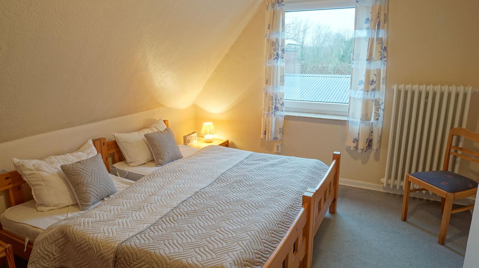 Schlafzimmer mit Bett, Nachttisch, Stuhl und Fenster mit Vorhängen.