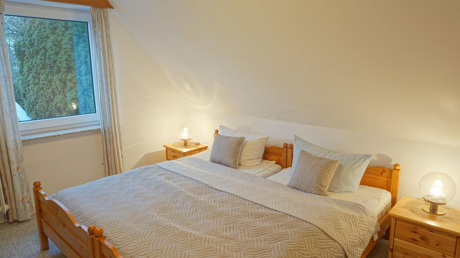 Schlafzimmer mit Holzbett, zwei Nachttischen und Fenster mit Blick auf Bäume.