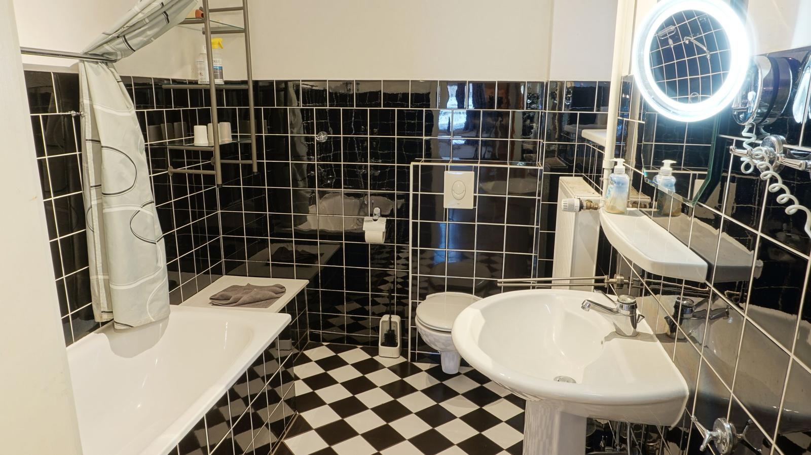 Badezimmer mit Badewanne, Waschbecken und Toilette. Schwarze Fliesen an den Wänden und schwarz-weißem Boden.