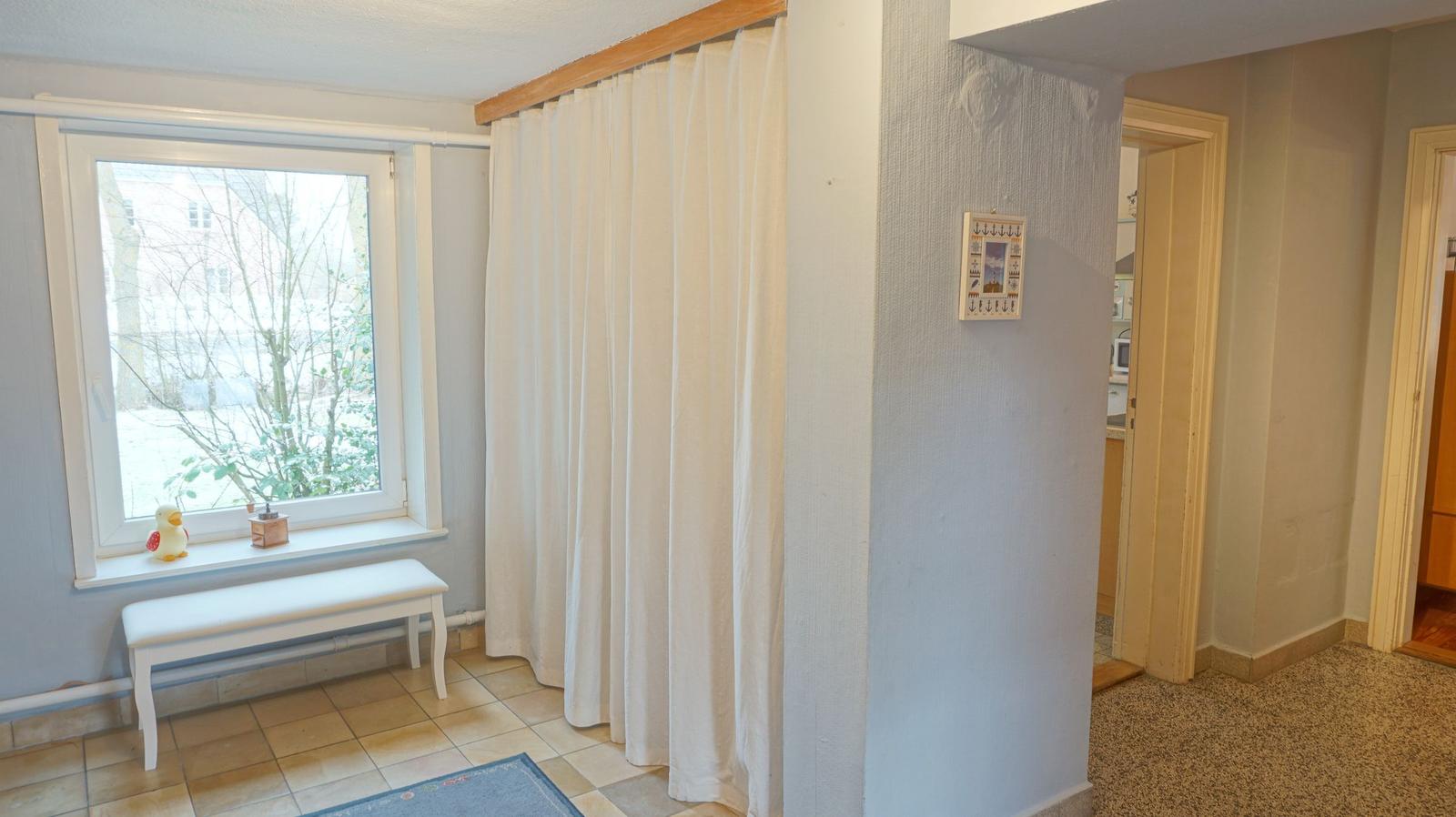 Raum mit Fenster, weiße Bank und Vorhang. Durchgang zu Küche und Schlafzimmer.
