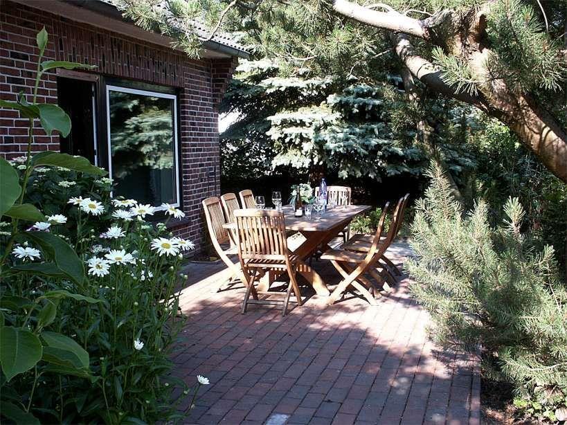 Terrasse mit Holztisch und Stühlen, umgeben von Pflanzen und Bäumen.