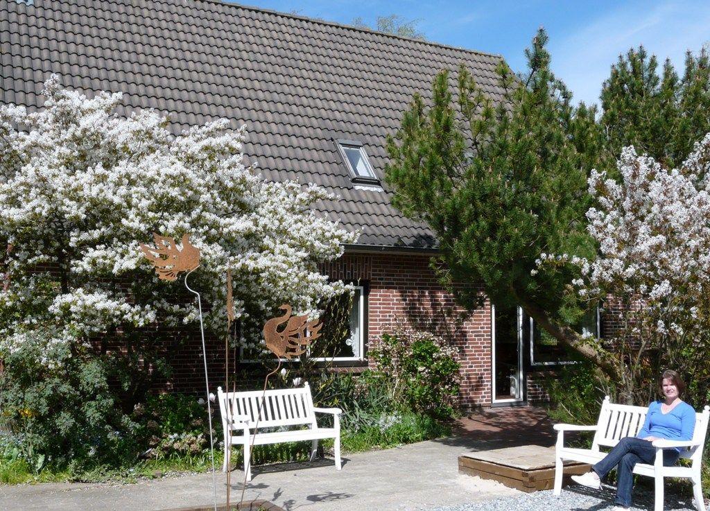 Haus mit Garten, weißen Bänken und blühenden Sträuchern unter blauem Himmel.