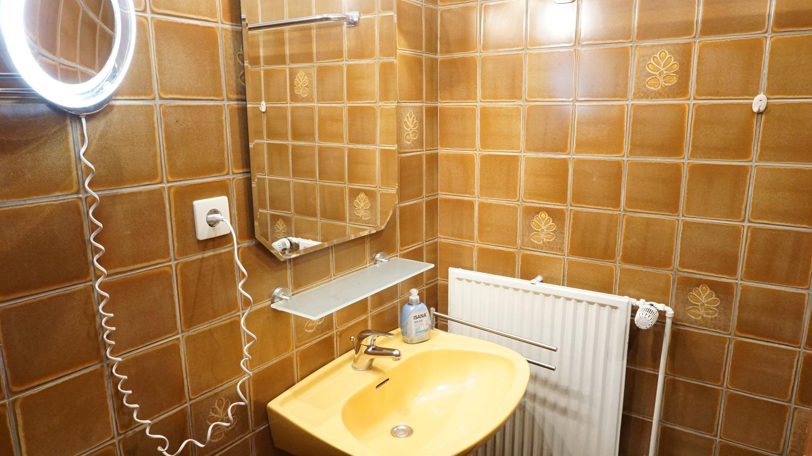 Badezimmer mit gelbem Waschbecken, Spiegel und Heizkörper an braunen Fliesenwänden.