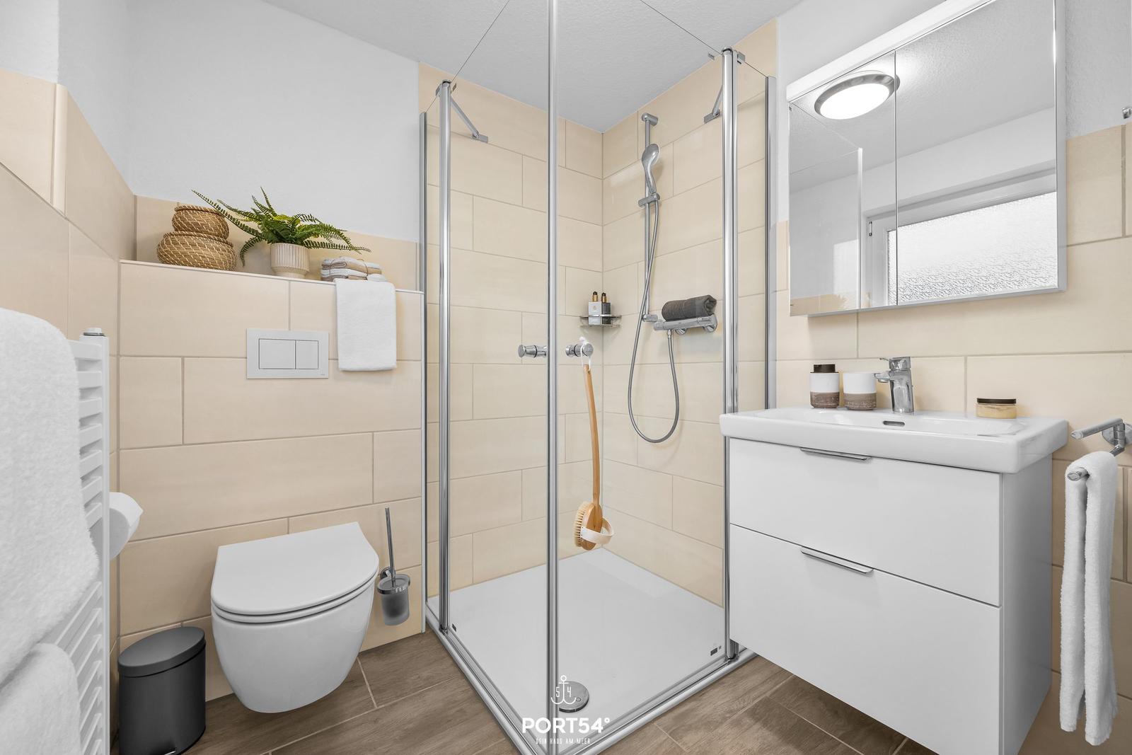 Badezimmer mit Dusche, Waschbecken und Toilette. Beleuchteter Spiegel und Handtuchhalter.