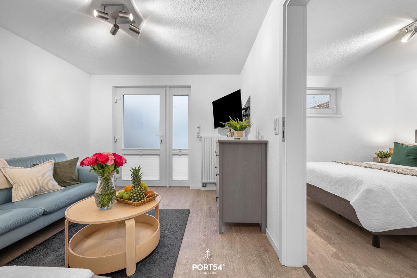 Wohnbereich mit Sofa, Couchtisch und Fernseher. Nebenraum mit Bett und Fenster.