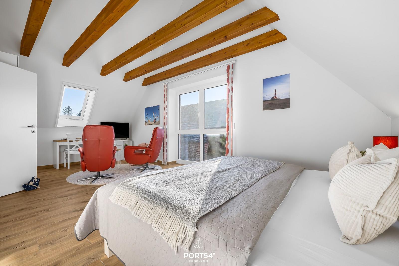 Schlafzimmer mit Dachschrägen, Holzbalken, Bett, zwei roten Sesseln und Fenstern.