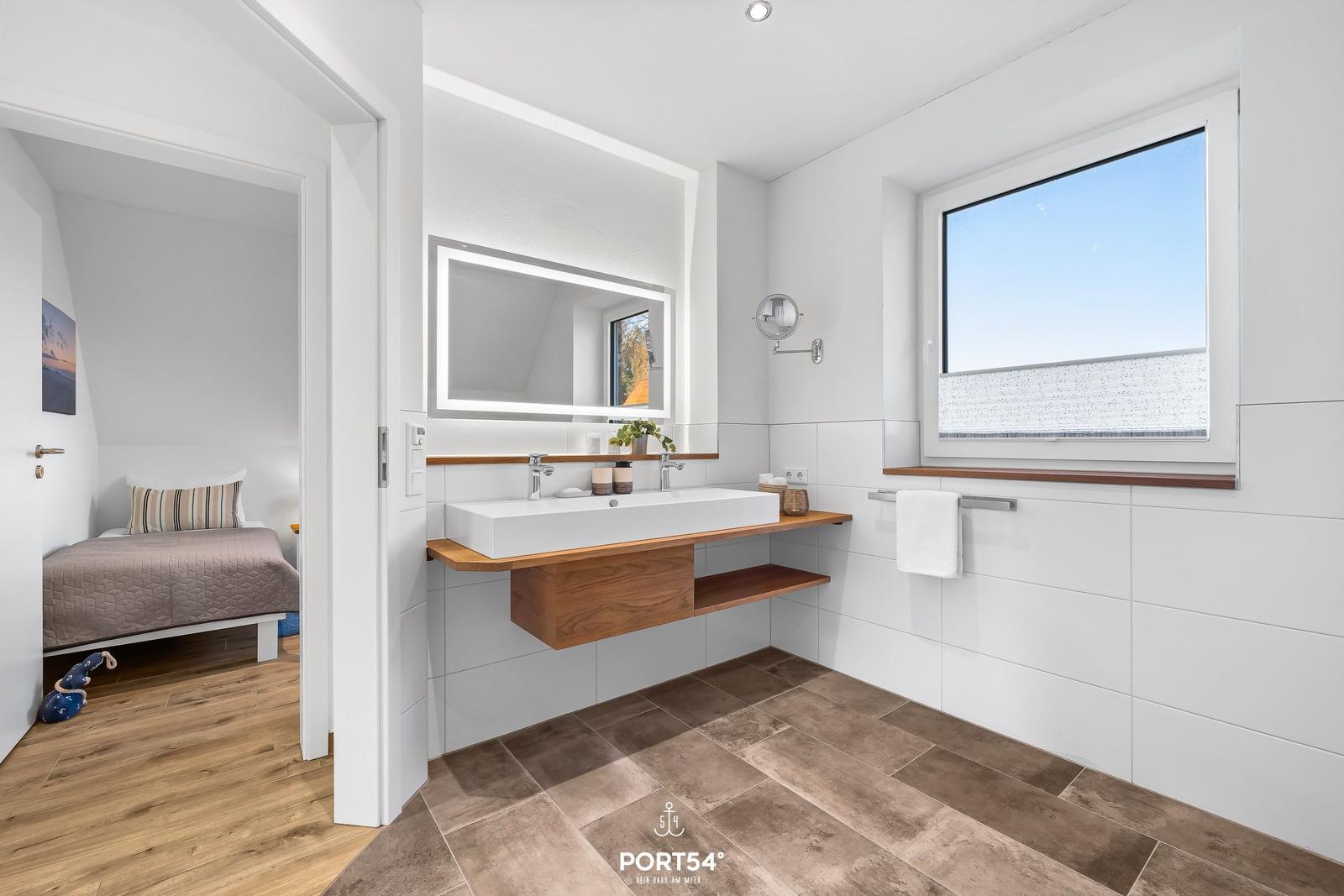 Badezimmer mit Waschbecken, Spiegel und Fenster. Durchgang zu Schlafzimmer mit Bett.