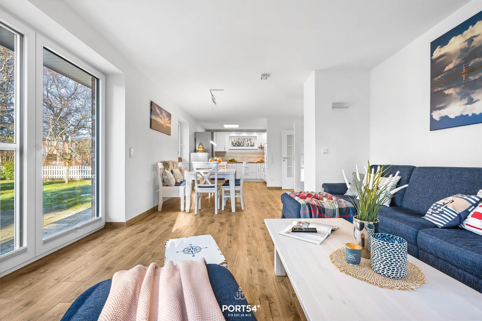 Wohn- und Essbereich mit Holzboden und großem Fenster. Sofa, Tisch und Küchenbereich sichtbar.