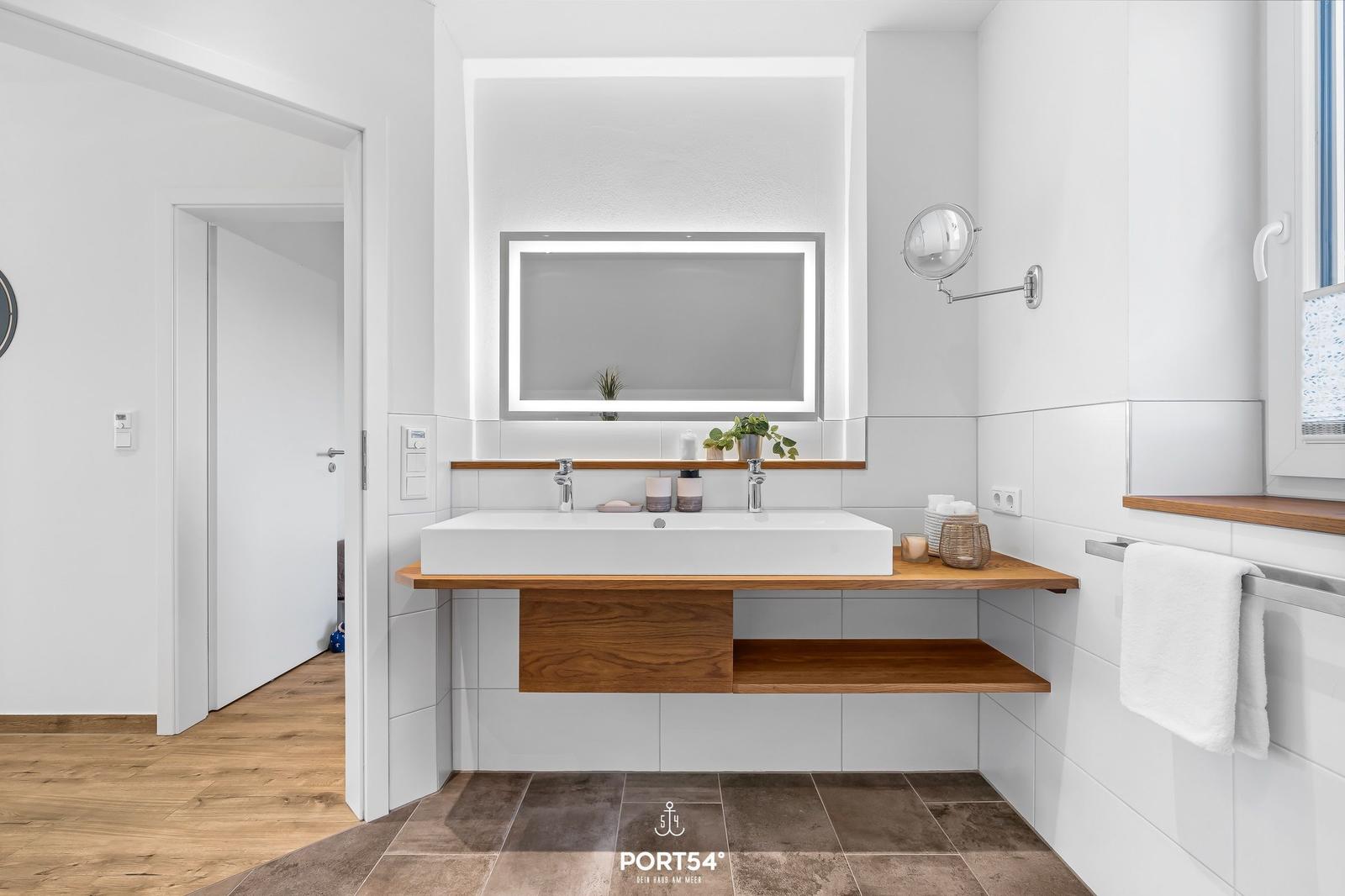 Bad mit Waschbecken, Spiegel und Holzregalen. Fenster und Handtuchhalter sichtbar.