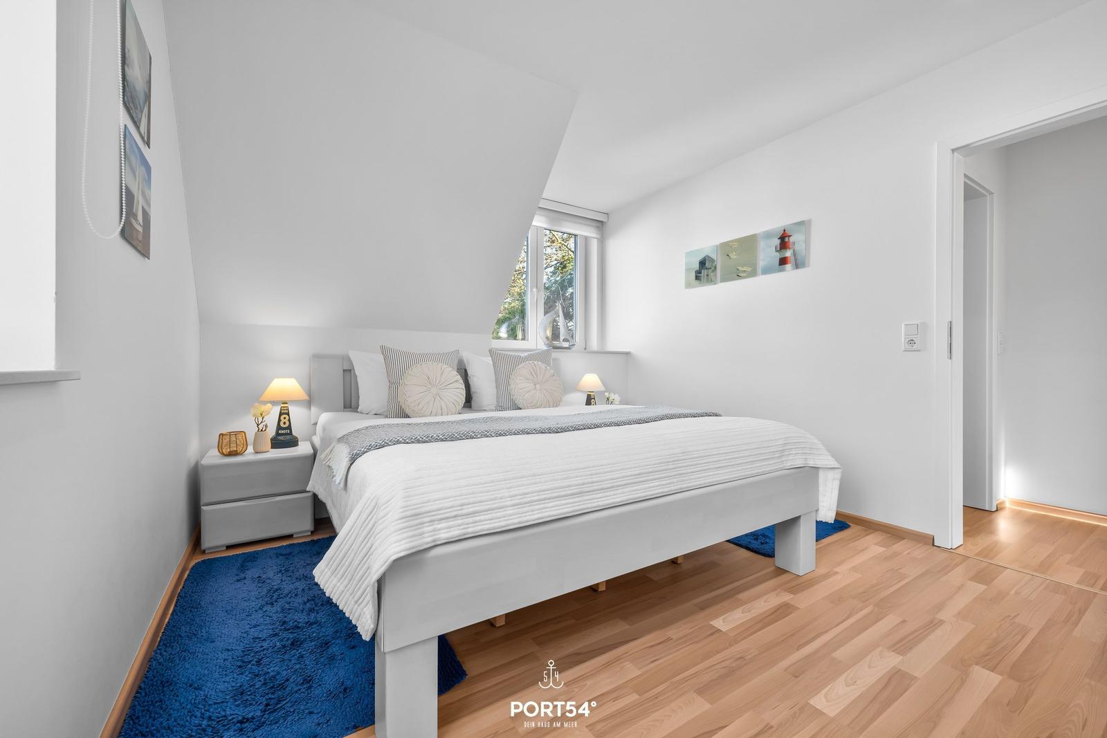 Schlafzimmer mit Bett, Nachttisch und Fenster. Blauer Teppich und Wandbilder.