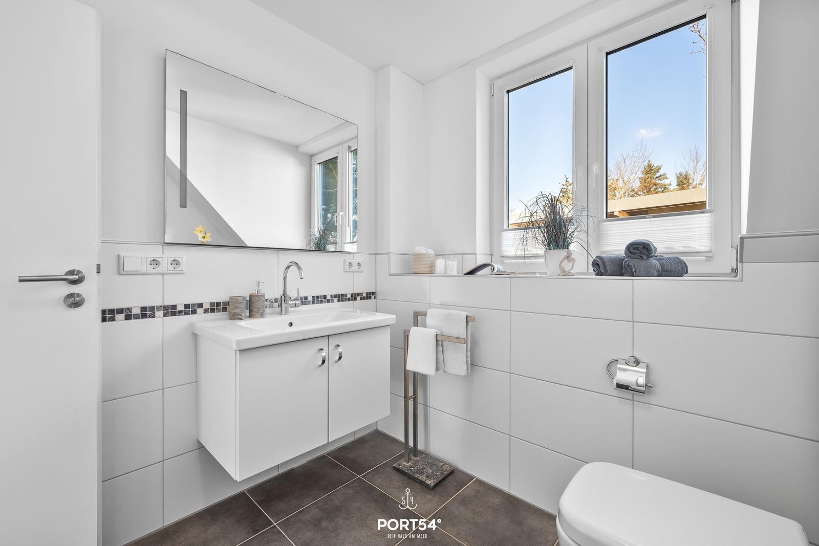 Badezimmer mit Waschbecken, Spiegel, Fenster und Toilette.