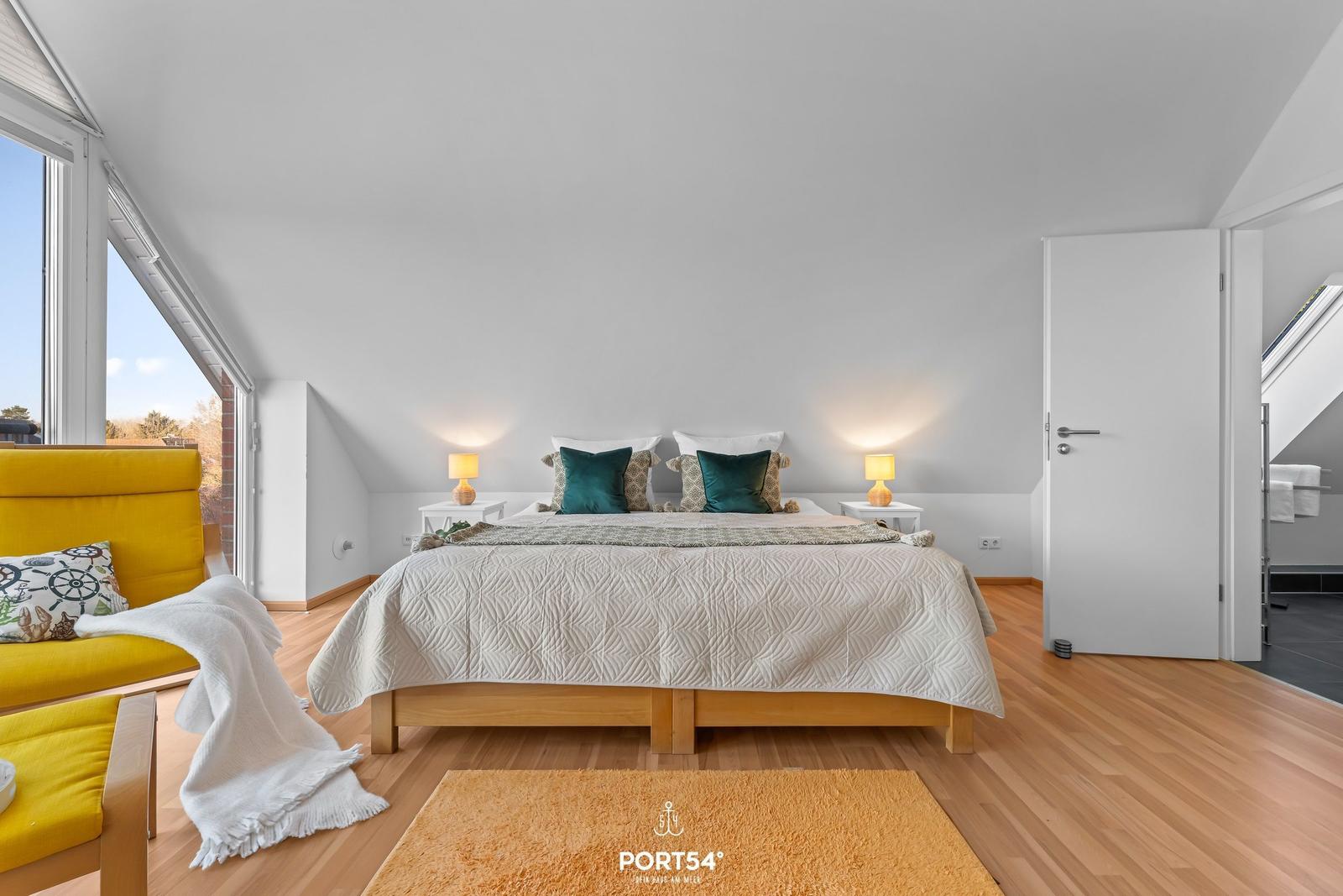 Schlafzimmer mit Bett, Nachttischen und gelbem Sofa. Holzboden und große Fenster.