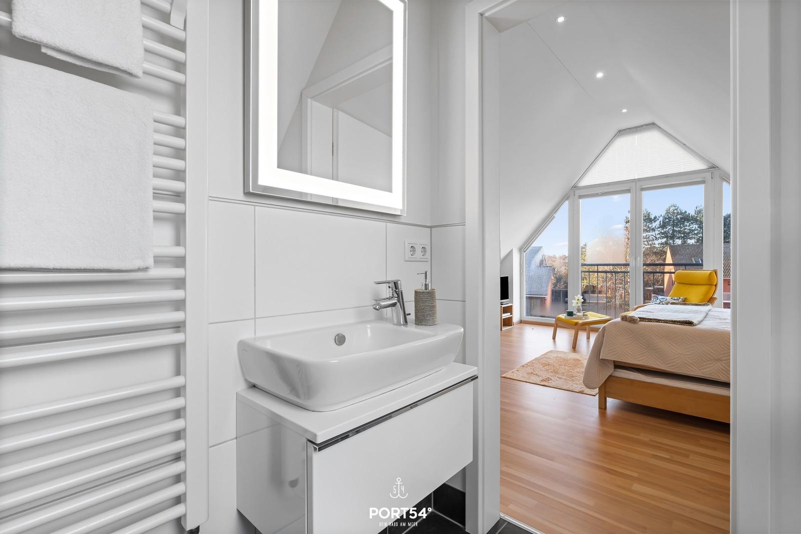 Badezimmer mit Waschbecken und Spiegel. Blick in Schlafzimmer mit Bett und Balkon.