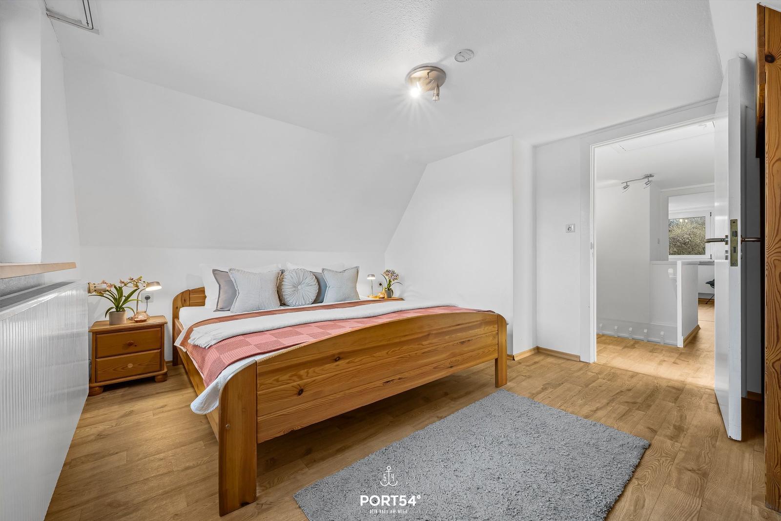 Schlafzimmer mit Holzbett, Nachttisch und Teppich. Tür führt in angrenzenden Raum.