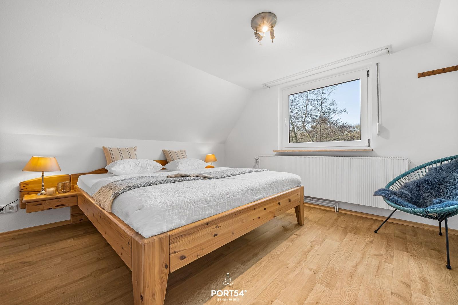 Schlafzimmer mit Holzbett, Nachttisch und Sessel. Fenster mit Blick auf Bäume.