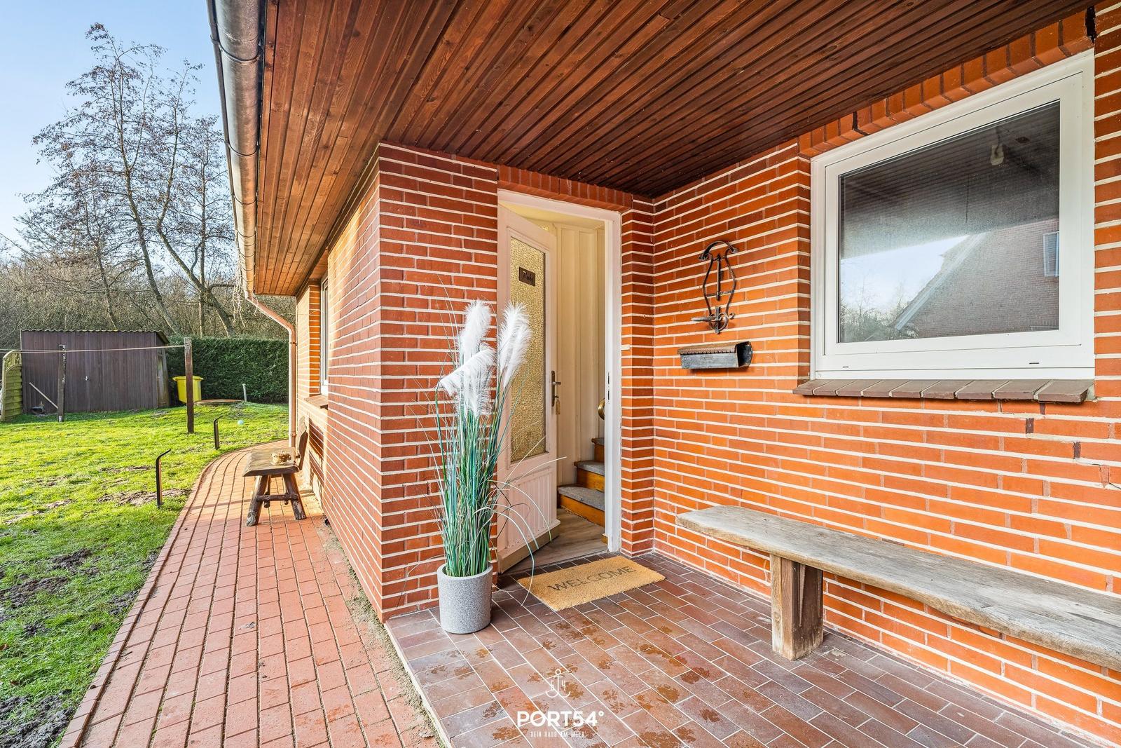 Eingangsbereich mit Holzveranda, Steinbank und Pflanze. Tür mit Glas und Briefkasten an der Wand.
