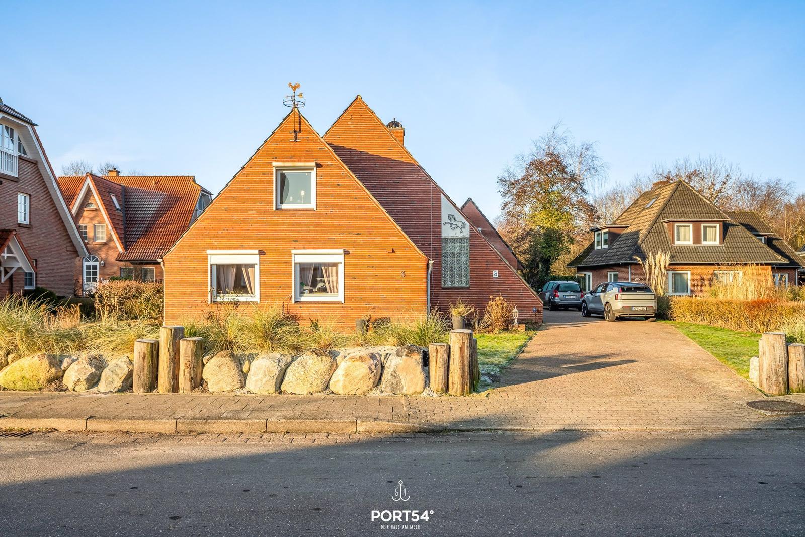 Rotes Backsteingebäude mit Dachgaube und Windschutz. Vorhof mit Stein- und Holzumrandung.
