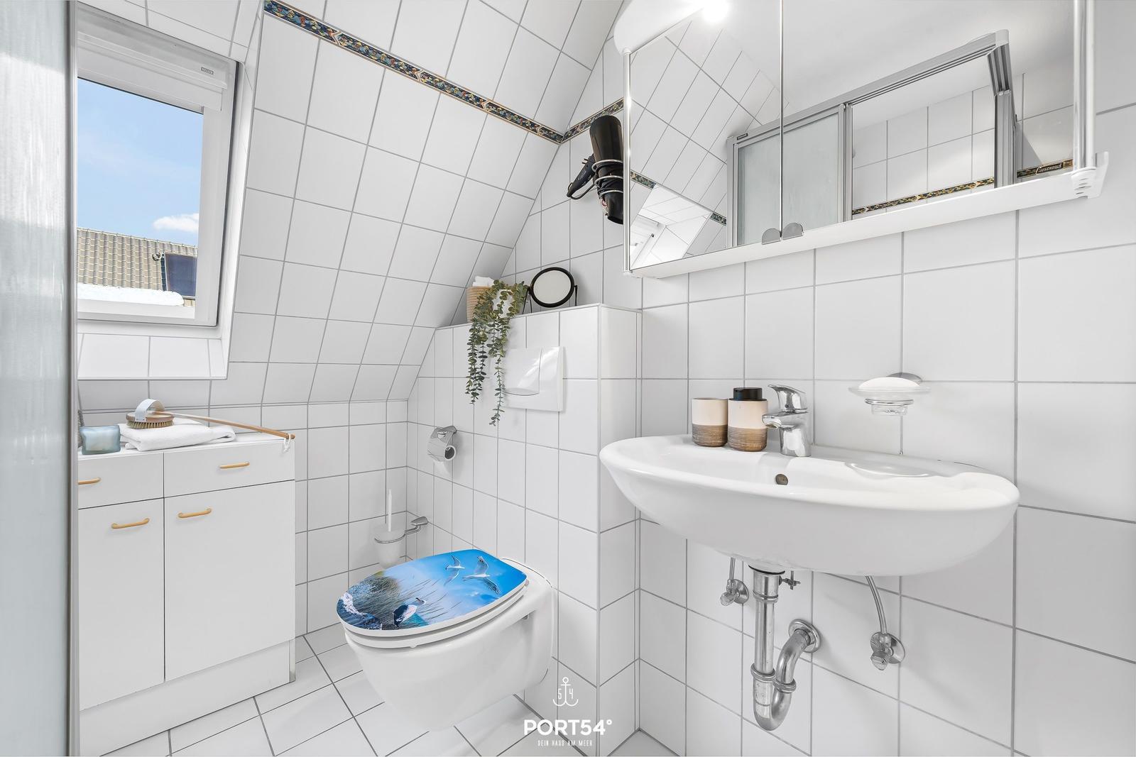 Badezimmer mit Toilette, Waschbecken und Fenster. Weiße Fliesen und Spiegelschrank.