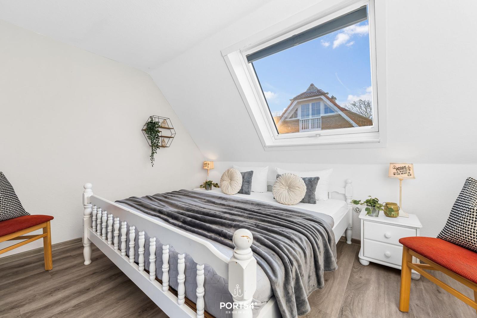 Schlafzimmer mit Dachfenster, weißem Bett und zwei Sitzbänken mit rotem Sitzkissen.