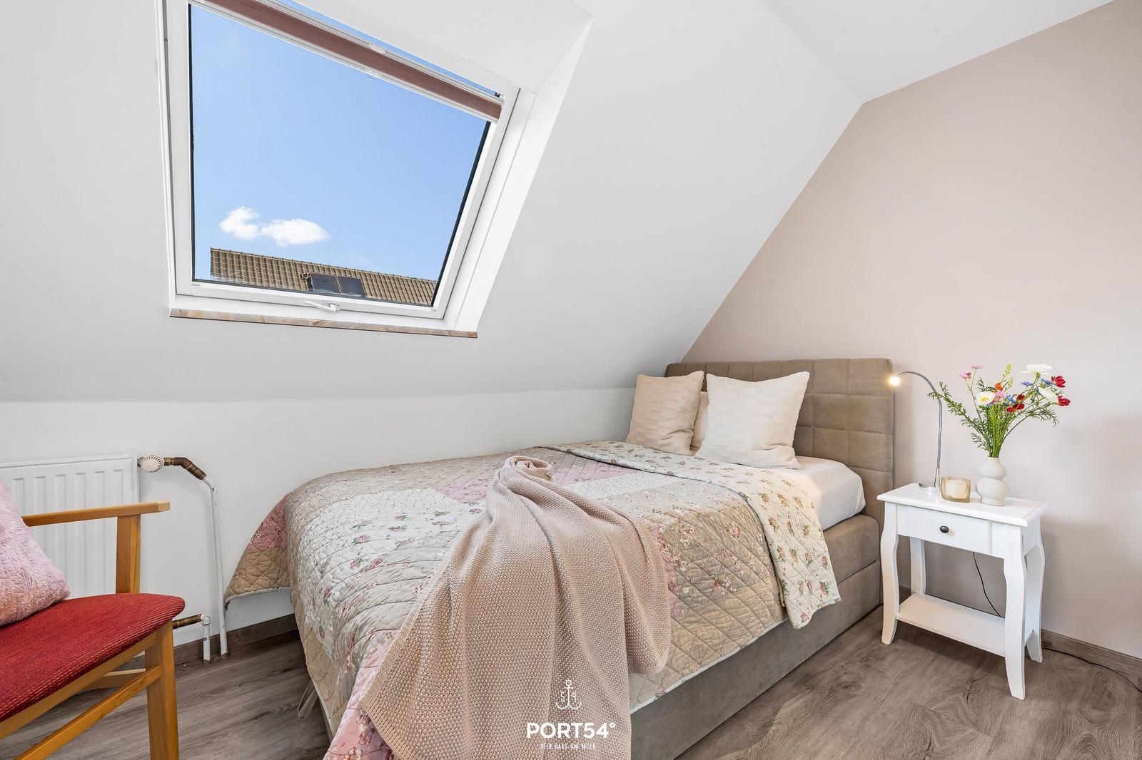 Schlafzimmer mit Dachschrägen, Bett, Nachttisch und Fenster mit Blick auf Dach.