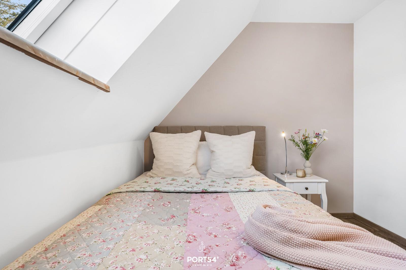 Schlafzimmer mit Dachschrägen, Bett mit Blumenbezug und Nachttisch mit Lampe.