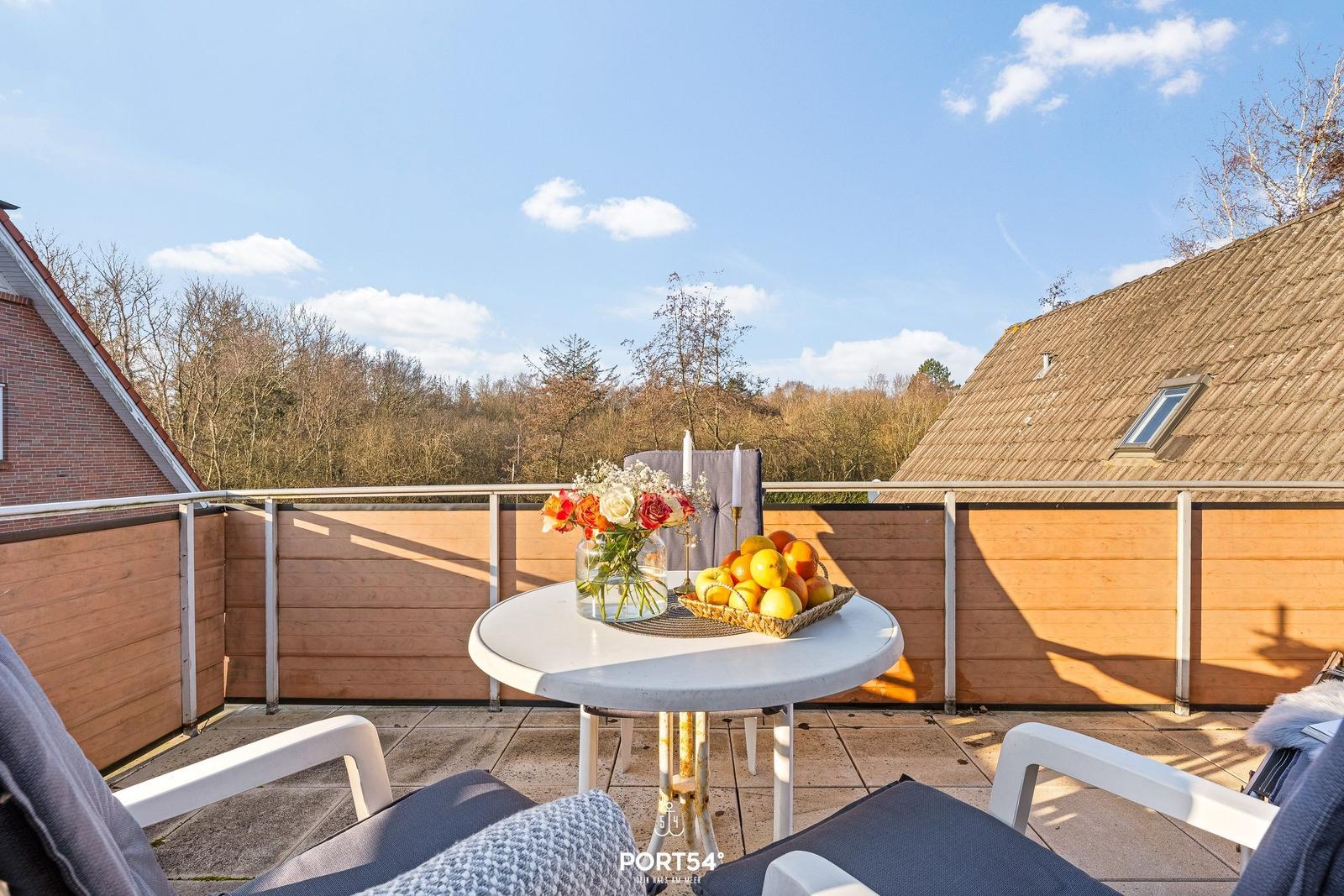Terrasse mit Tisch, Stühlen und Obstschale. Blick auf Wald und Häuser.