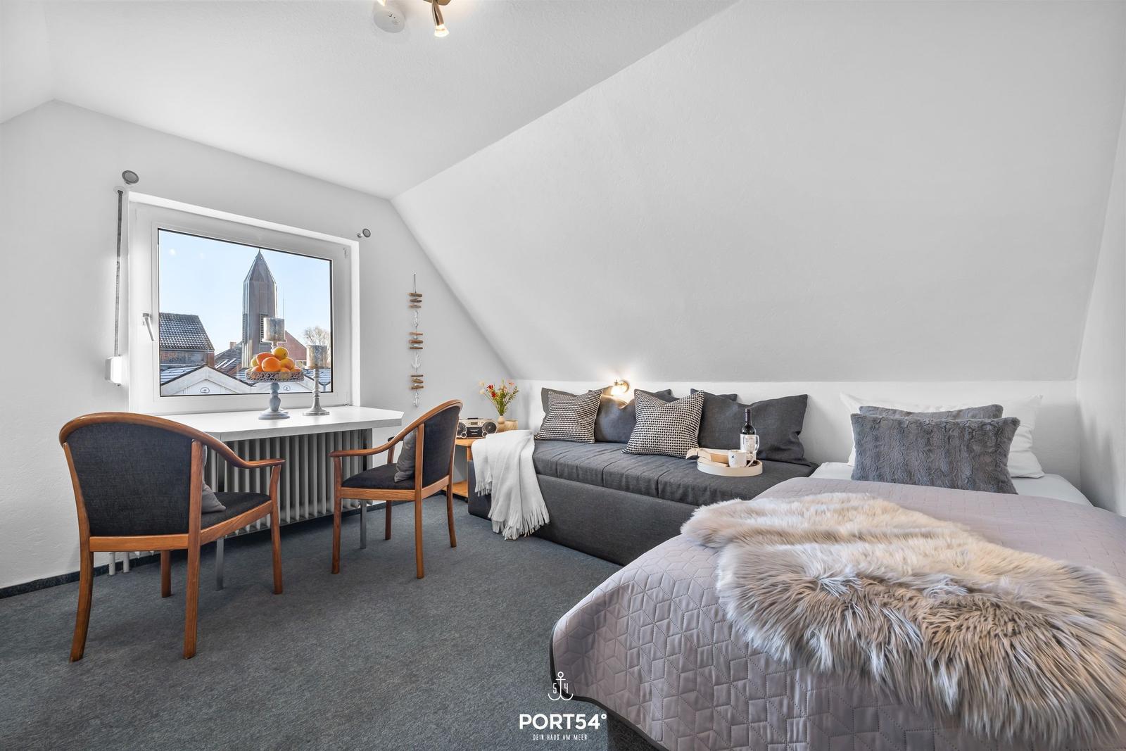 Schlafzimmer mit Dachschrägen, Bett, Sofa und Fenster mit Blick auf Kirche.