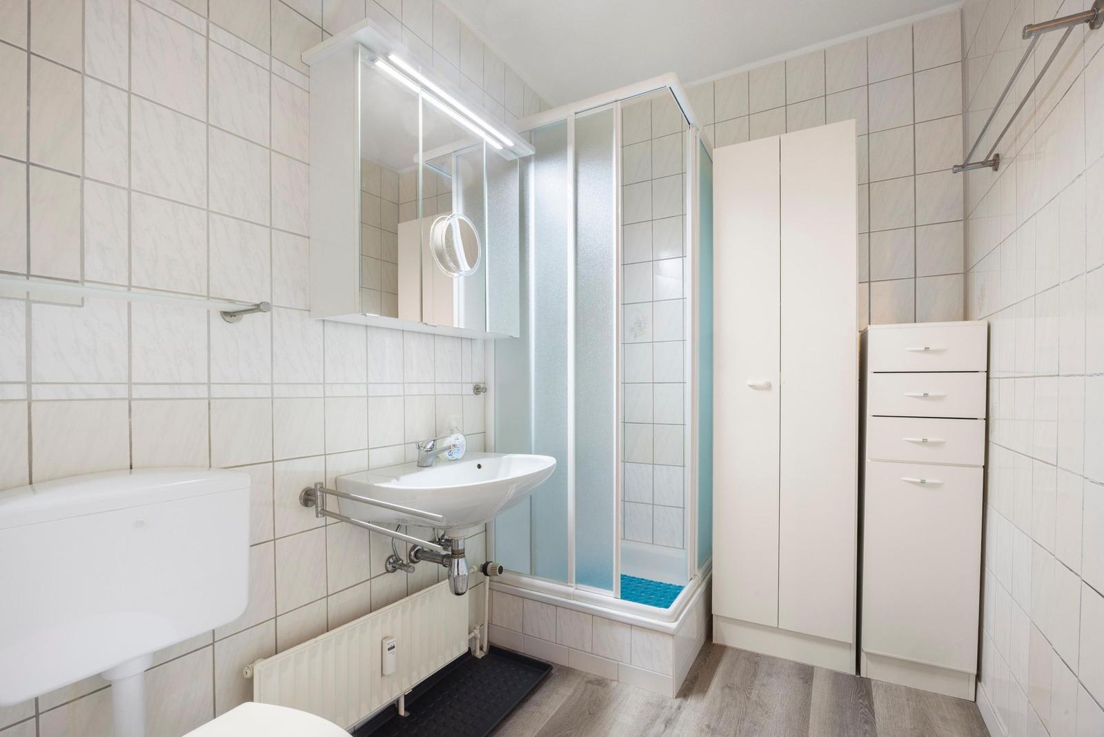 Badezimmer mit Dusche, Waschbecken und Toilette. Weiße Fliesen und Schränke.