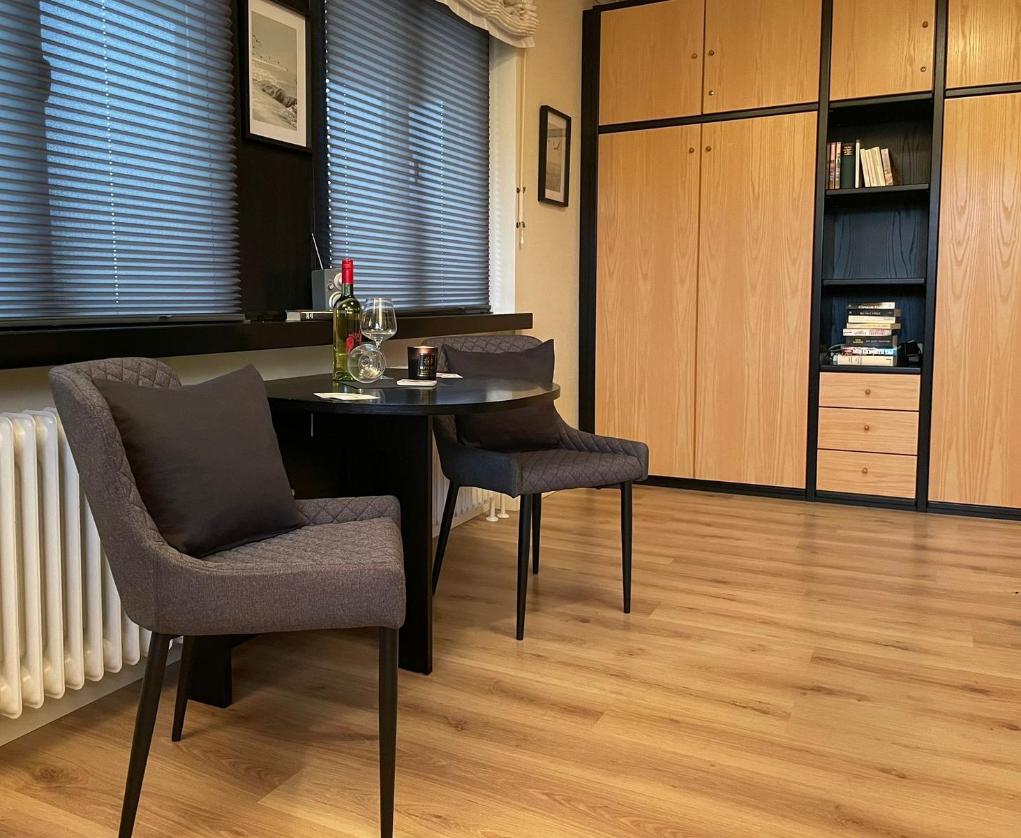 Zimmer mit Tisch, zwei Stühlen, Bücherregal und Fenster mit Jalousien.