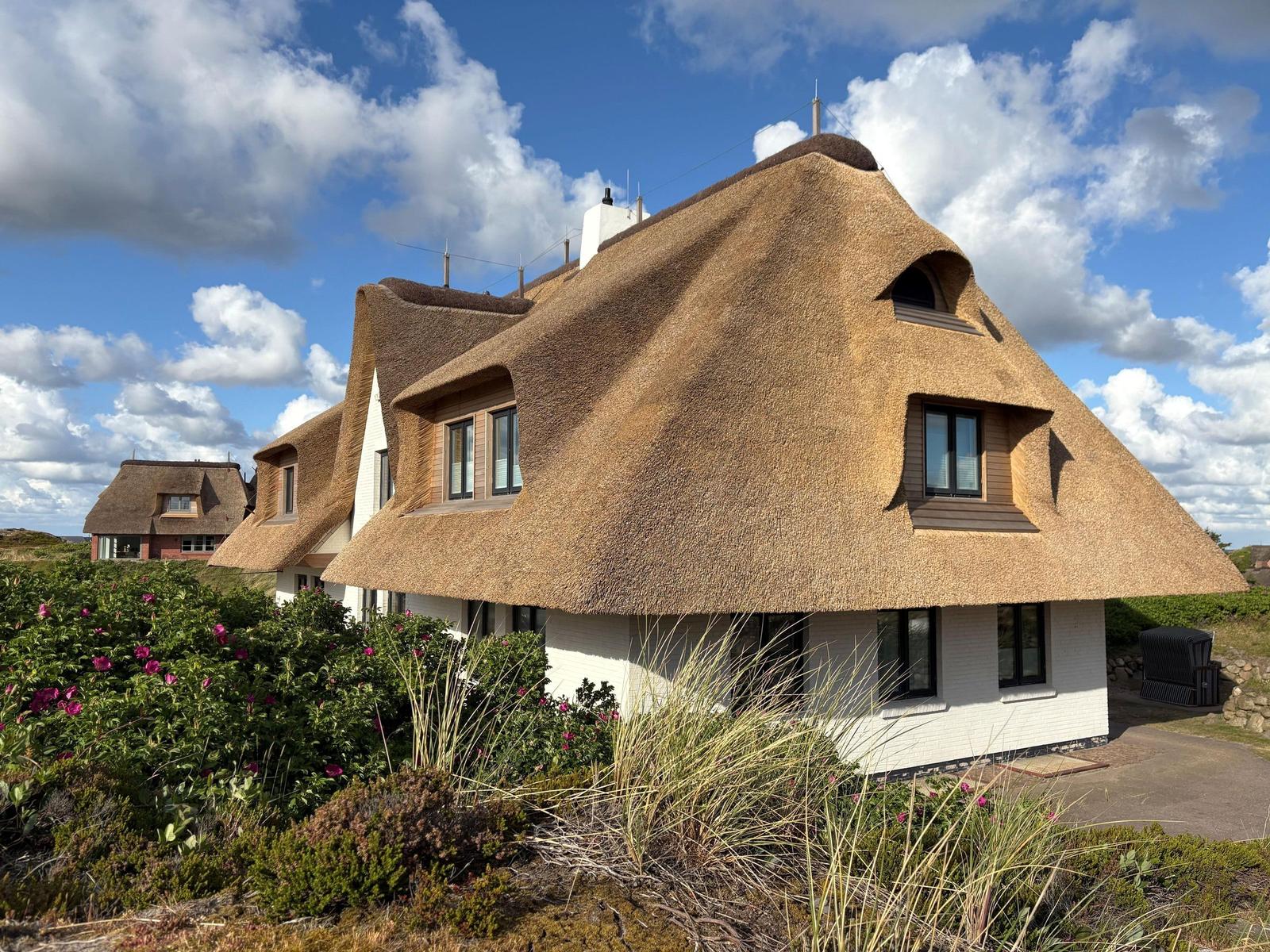 Haus mit strohgedecktem Dach und weißen Wänden, umgeben von Grünflächen und Blumen.