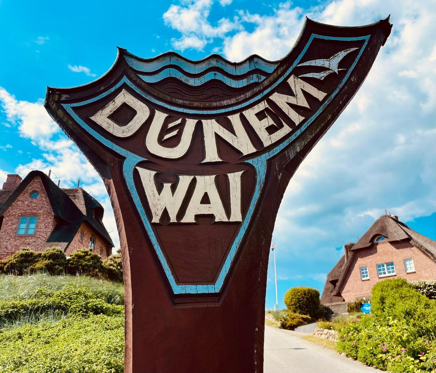 Willkommensschild für Dunenweg mit traditionellen Häusern im Hintergrund.