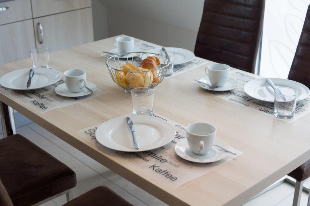 Esstisch mit Platz für vier Personen, Tassen, Tellern und Brötchen.