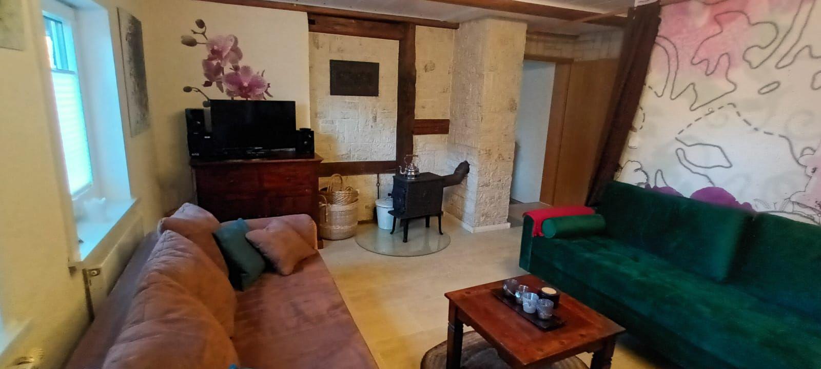 Wohnzimmer mit grünem Sofa, Holztisch und Kaminofen. Wandbild und Fenster sichtbar.