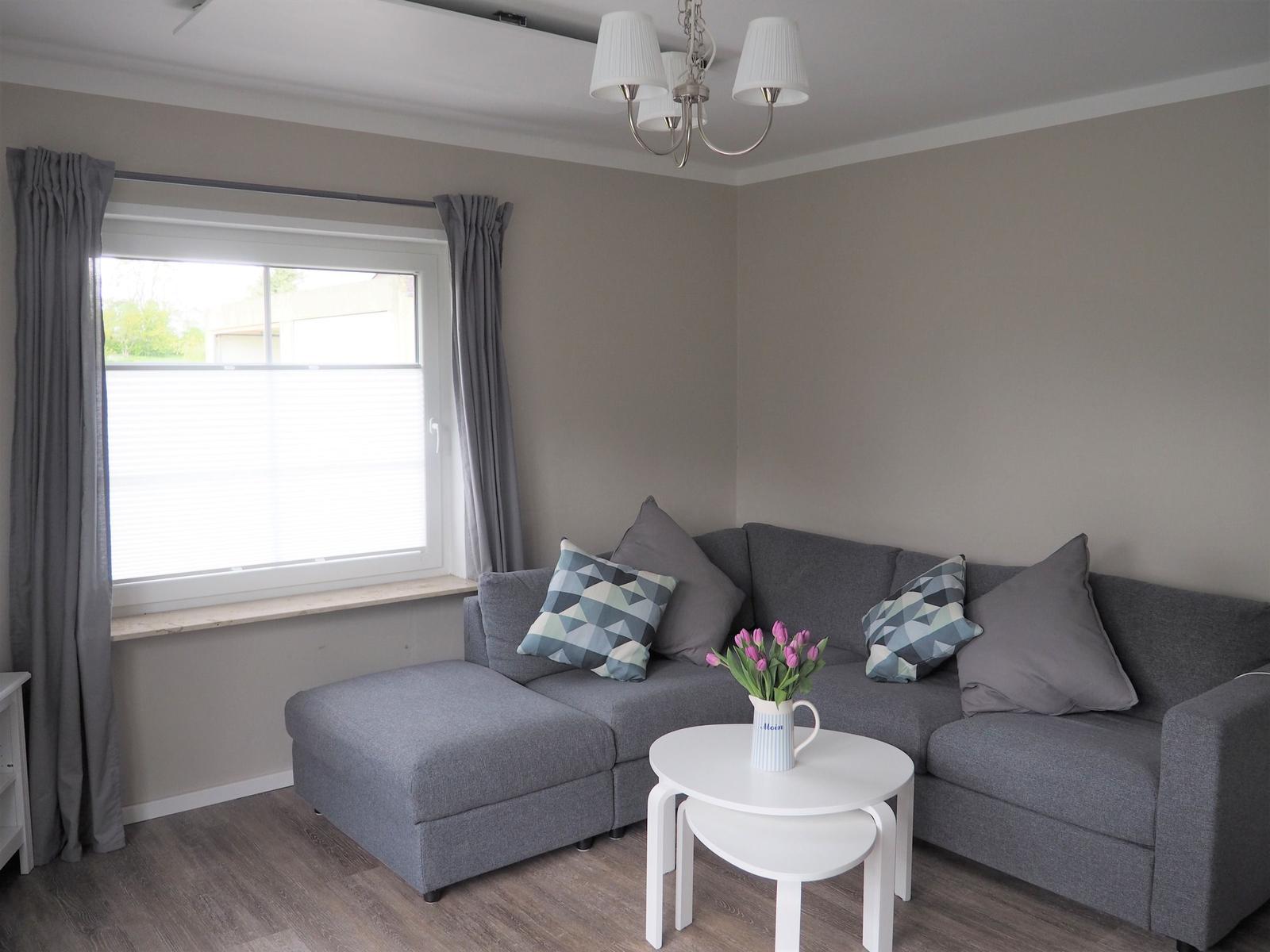 Ein Wohnzimmer mit grauem Sofa, weißem Tisch und Fenster mit Jalousie.